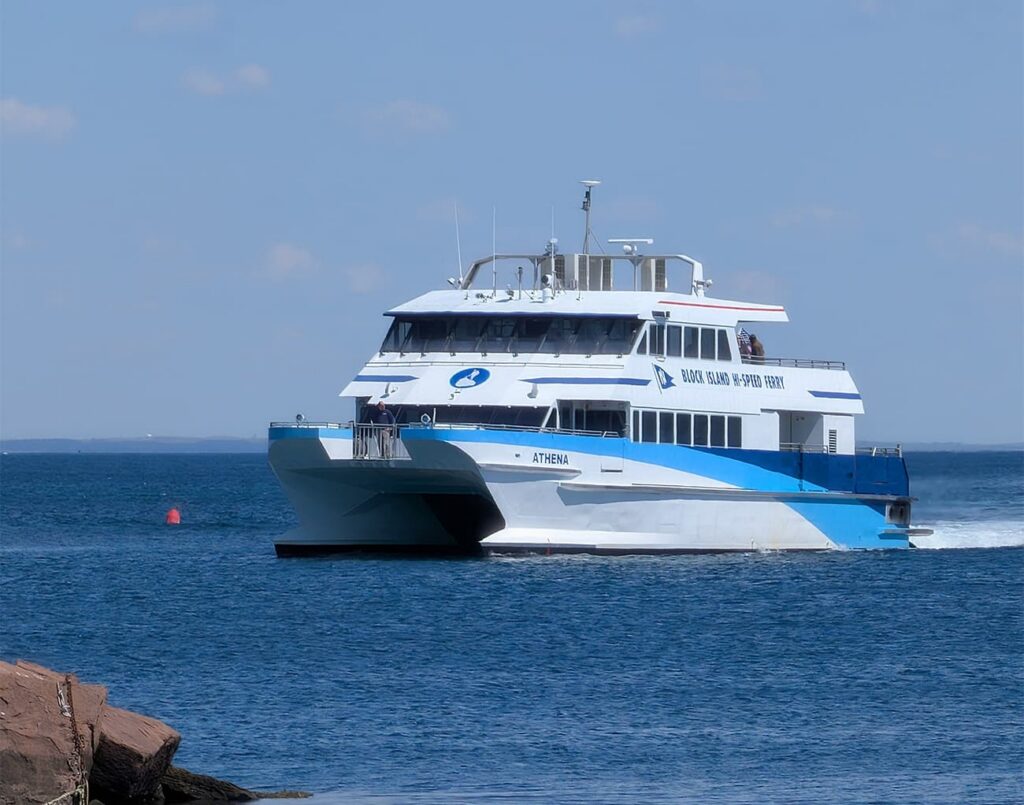 A white and blue Block Island Ferry named "ATHENA" sails toward Block Island on calm water, with people on deck; rocks and a red buoy in front.