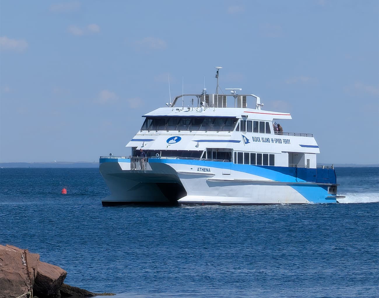 A white and blue Block Island Ferry named "ATHENA" sails toward Block Island on calm water, with people on deck; rocks and a red buoy in front.